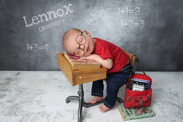 Newborn in a red shirt and jeans and glasses sitting on a small desk with a chalkboard background displaying mathematical equations.