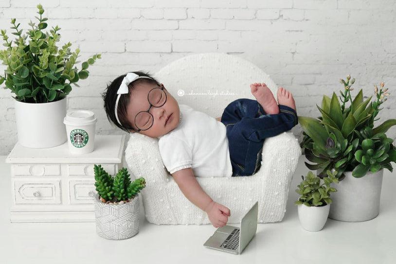 Baby in a white t-shirt and jeans with a laptop on a white surface surrounded by potted plants.